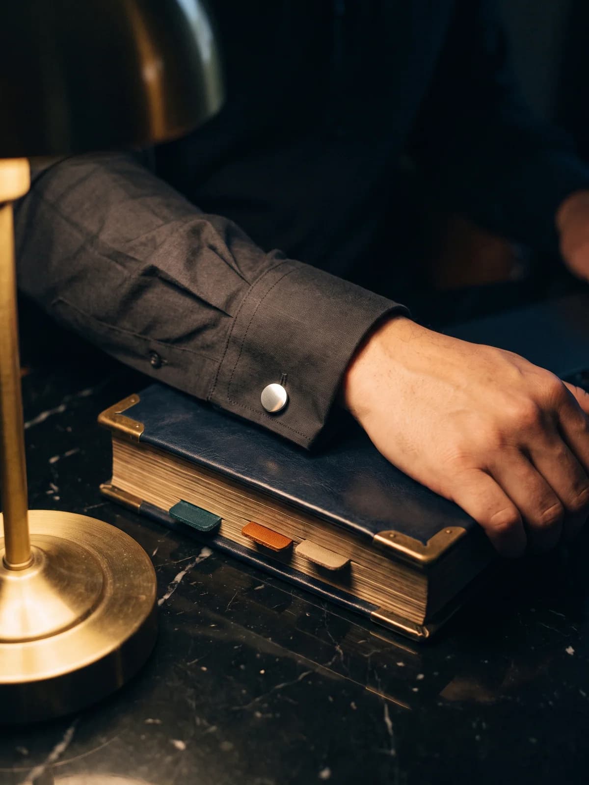 A hand resting on a closed navy leather ledger with brass corners and teal, amber and sand index tabs, under a brass desk lamp